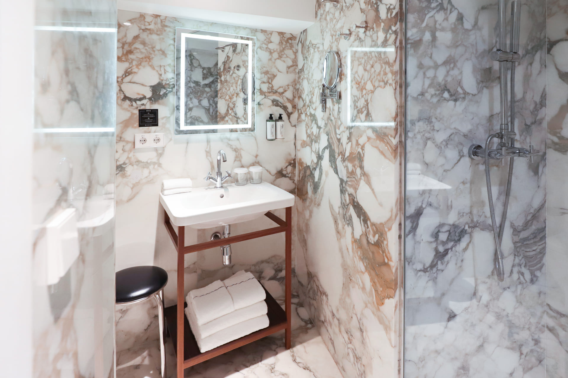 Baño de mármol con lavabo blanco sobre mueble de madera y ducha de vidrio