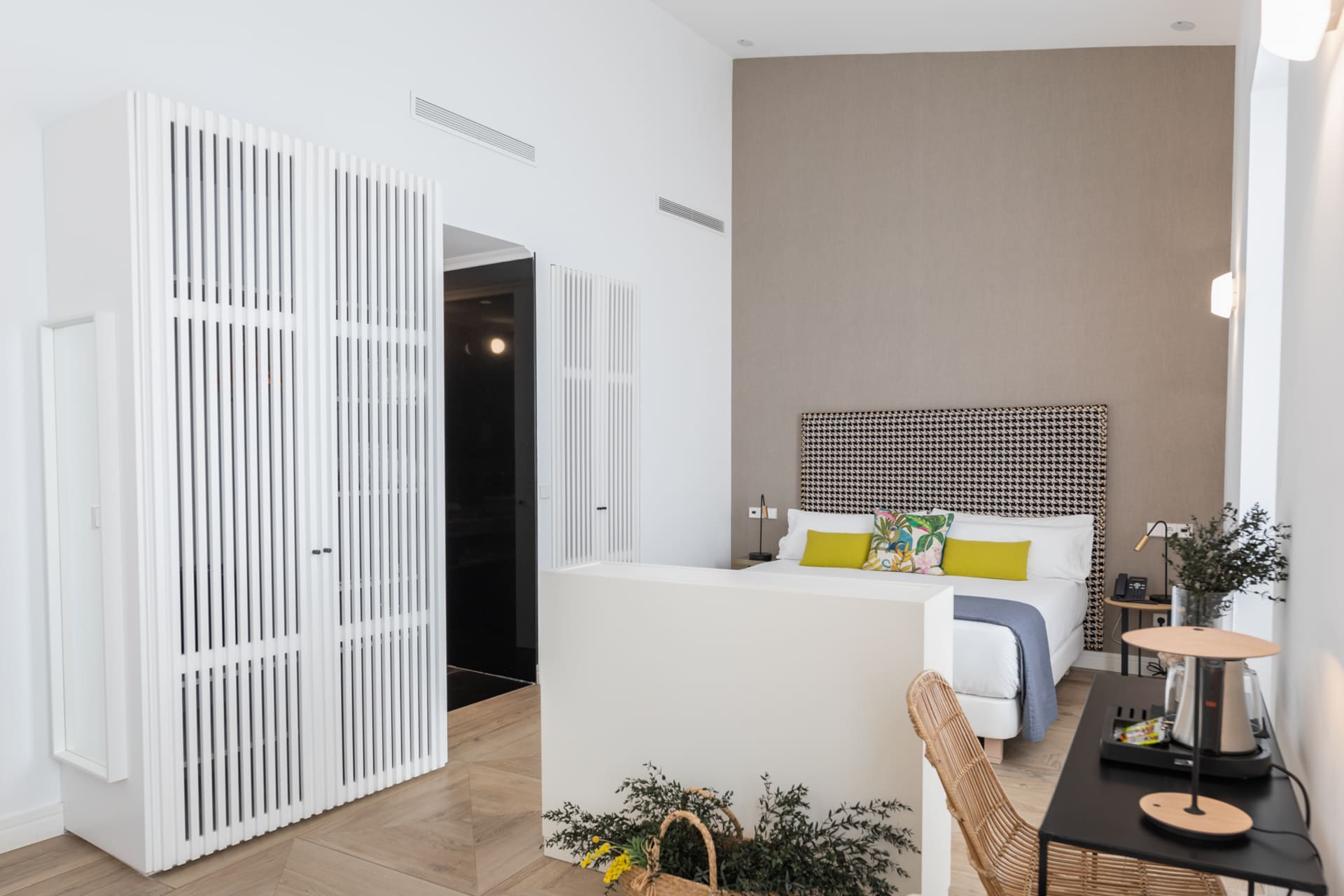 Habitación de hotel con cama y cabecero estampado, cojines verdes y zona de escritorio.