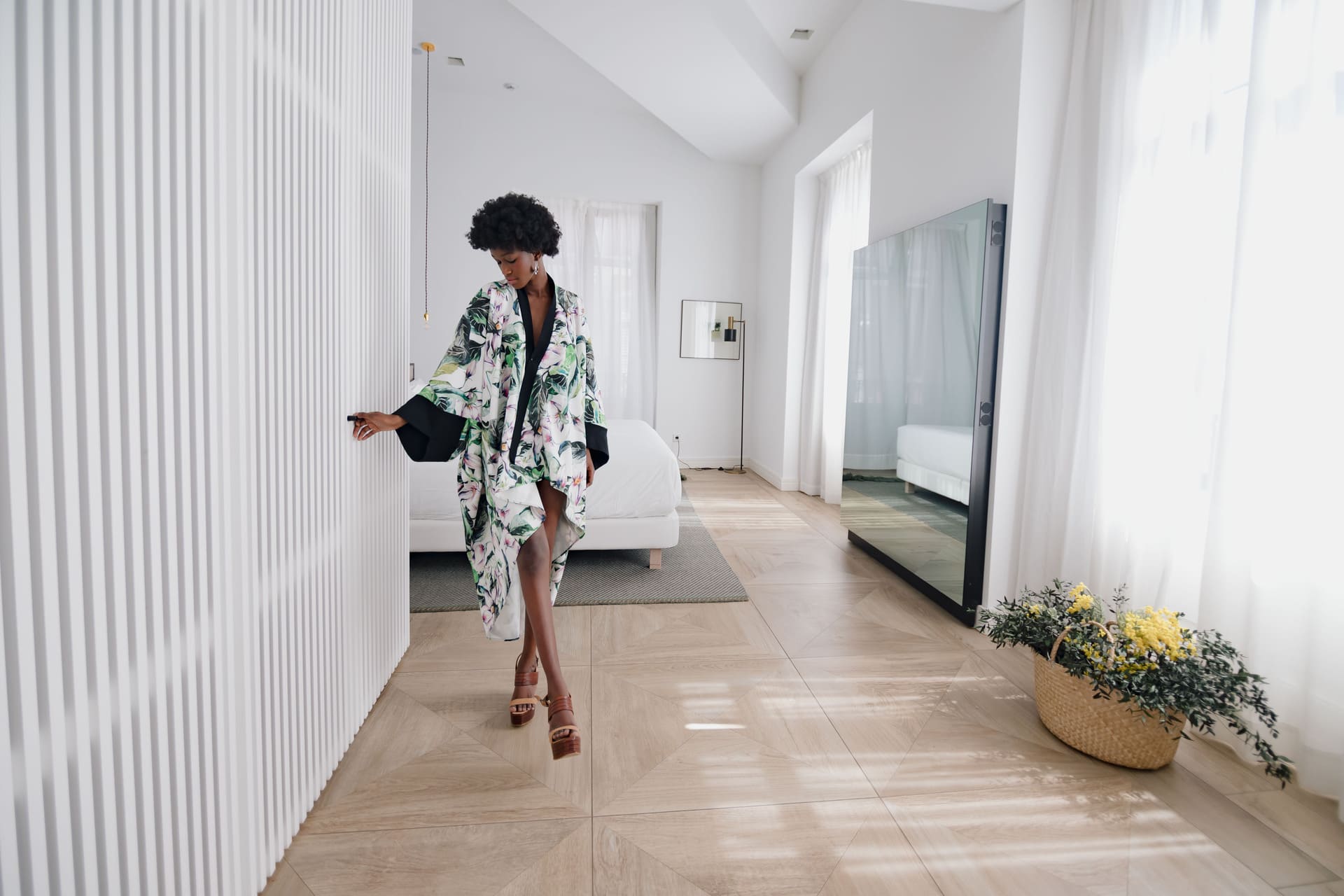 Mujer con kimono floral camina por pasillo de hotel, junto a cama blanca y espejo