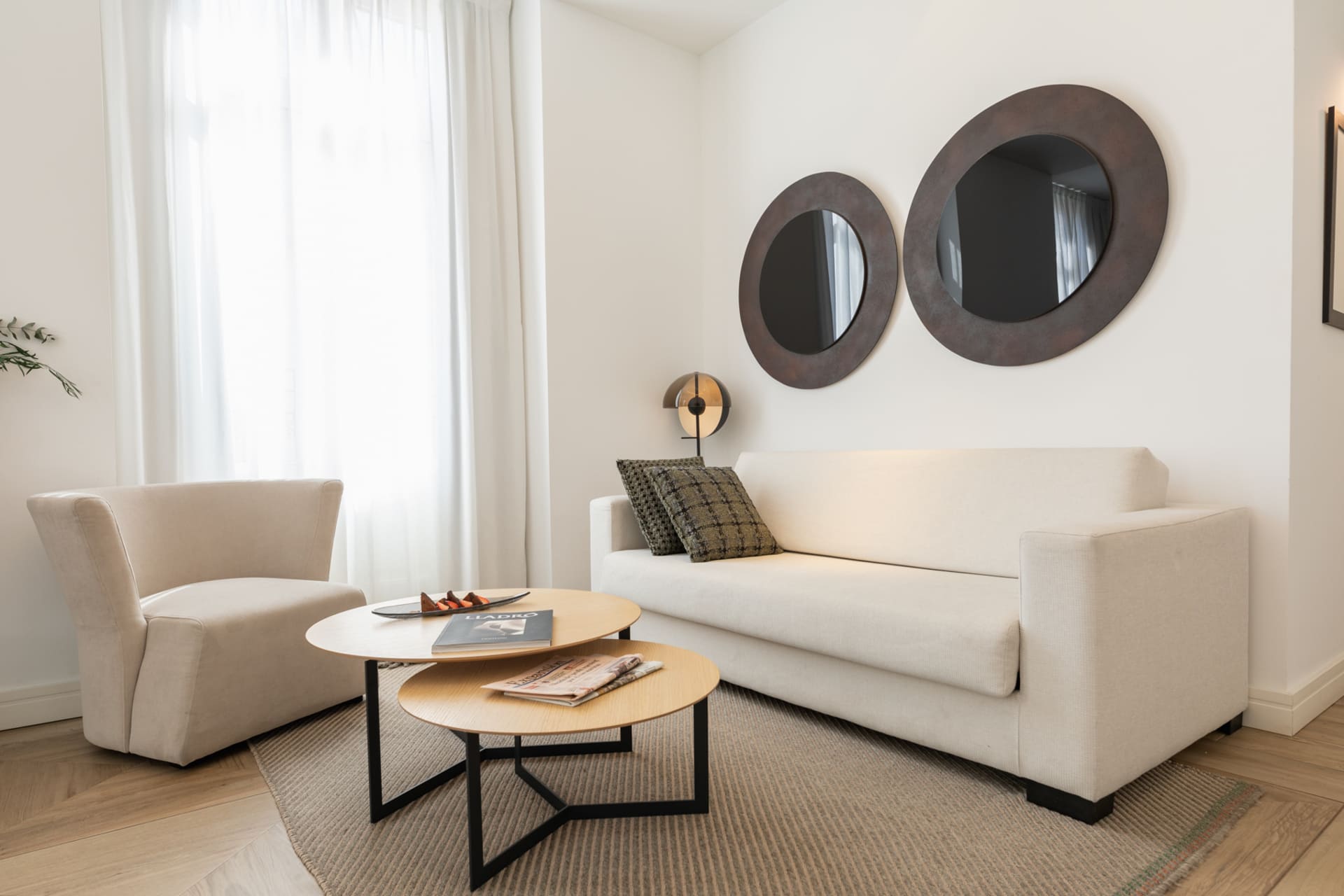 Sala de estar de habitación de hotel con sofá beige, sillón y dos espejos circulares en la pared