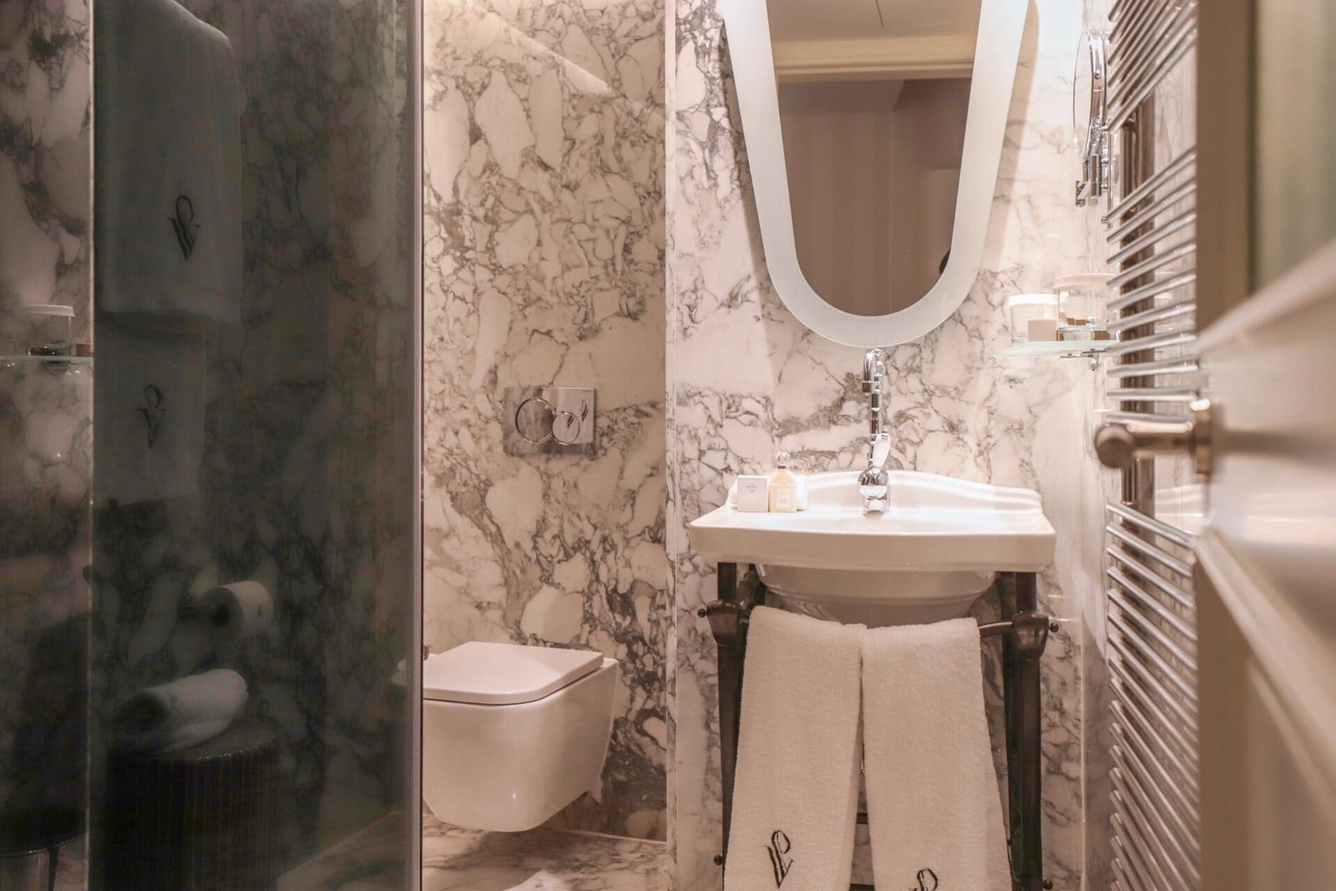 Baño de hotel con lavabo blanco, espejo ovalado, toallas colgando y paredes de mármol