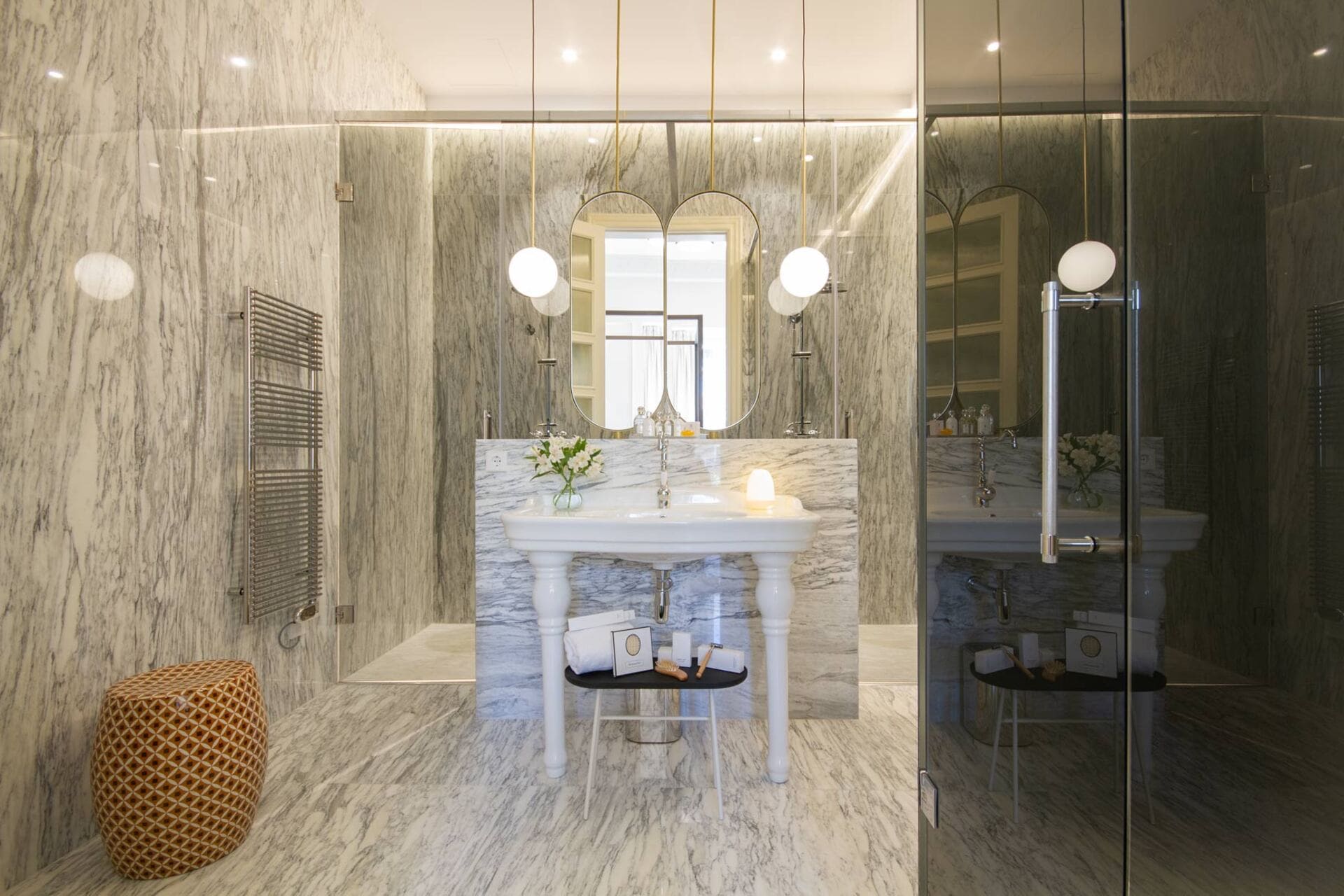 Baño de mármol con lavabo de pedestal blanco, espejo ovalado y ducha de vidrio; lámparas colgantes.
