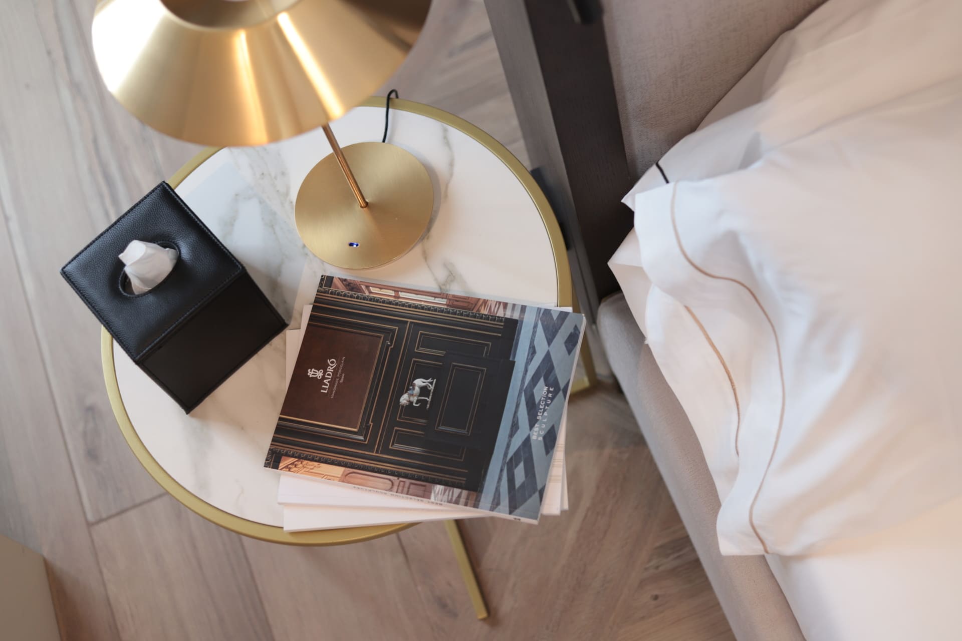 Mesa de noche redonda con superficie de mármol, lámpara dorada, caja de pañuelos y folleto junto a la cama.