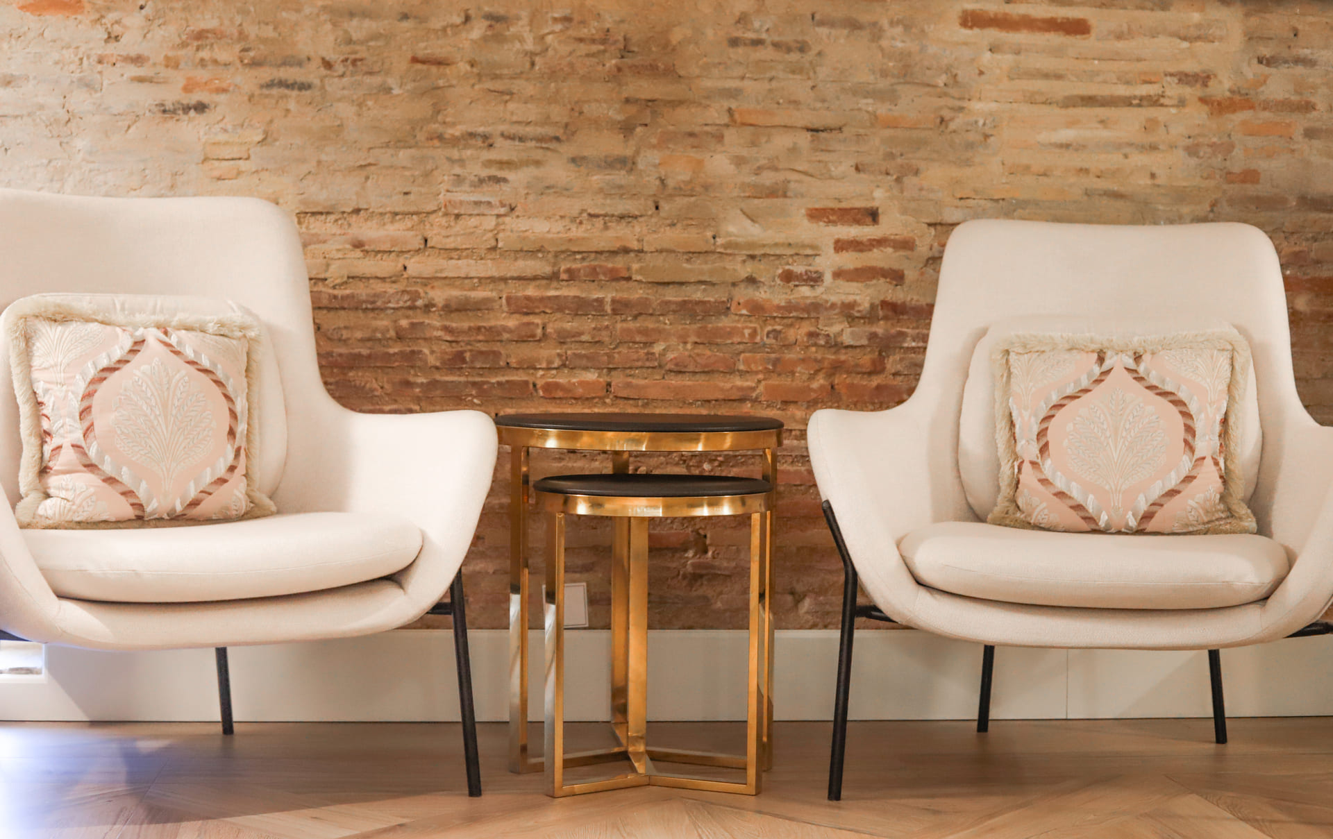 Dos sillones blancos con cojines decorativos entre ellos, junto a una mesa auxiliar dorada frente a una pared de ladrillo.