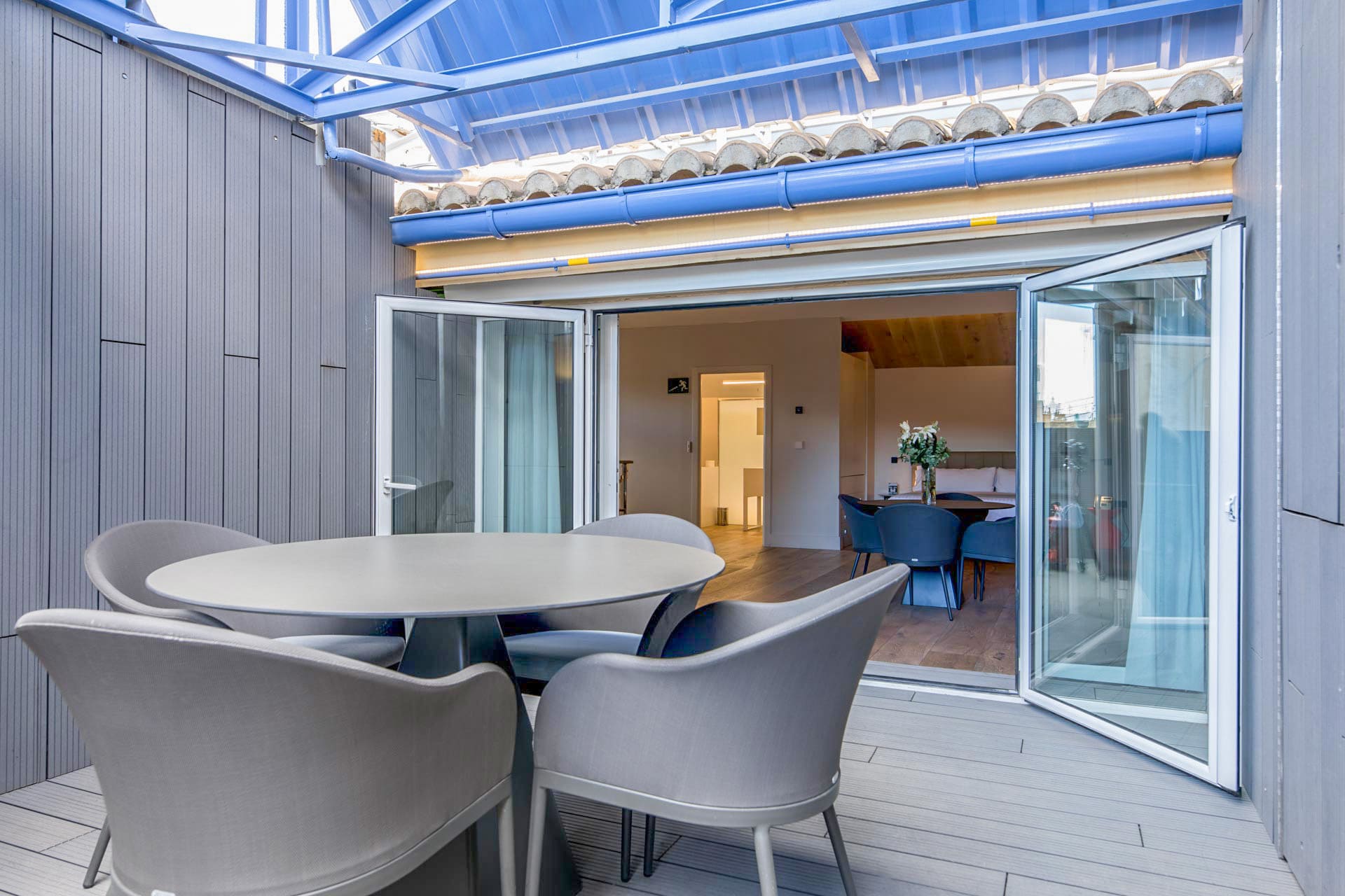 Terraza exterior con mesa redonda y sillas grises, puertas correderas abiertas a la sala de comedor.