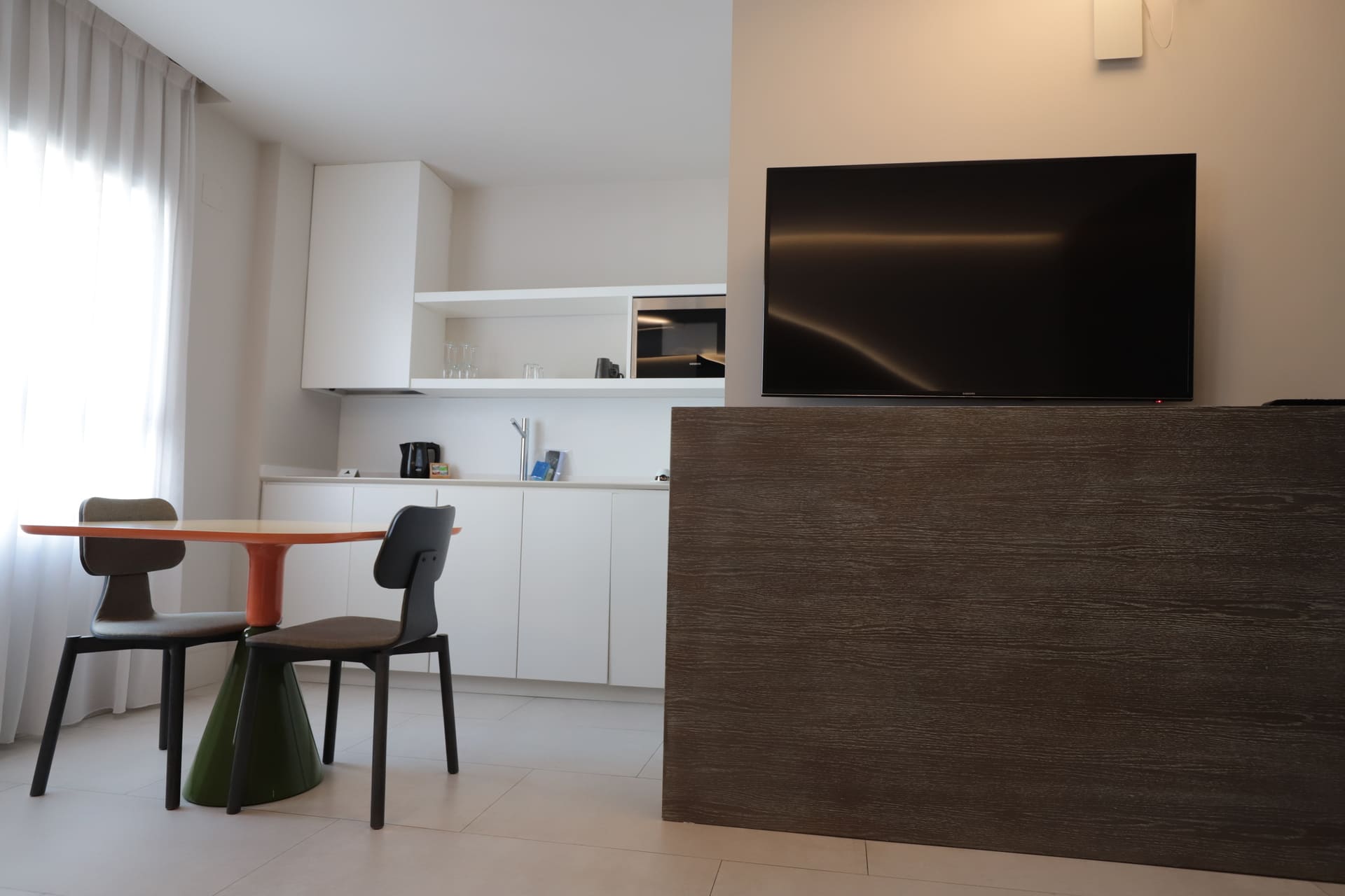 Sala de estar con cocina integrada, mesa redonda naranja y dos sillas, TV frente a panel de madera