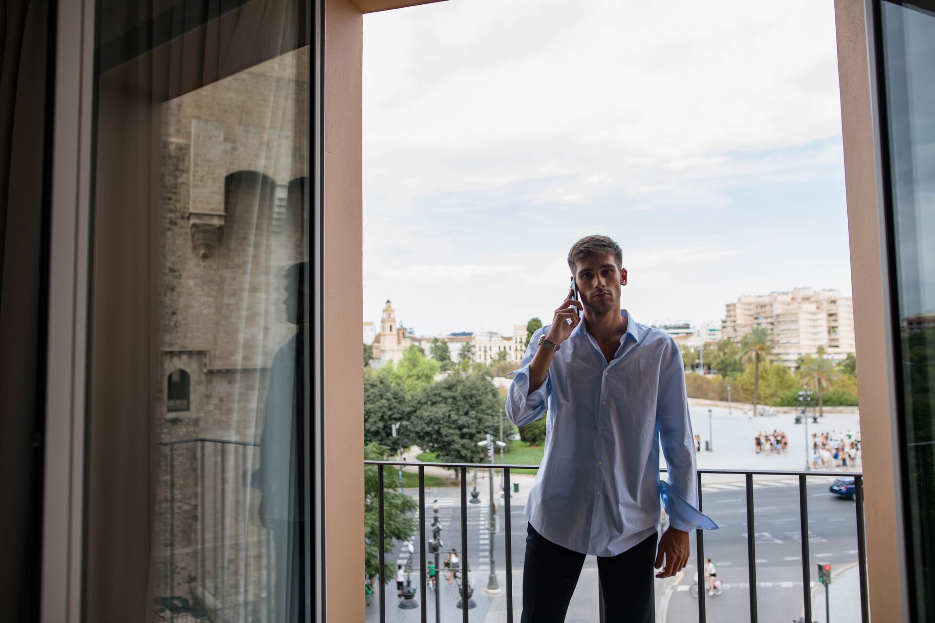 Hombre con camisa azul habla por teléfono en balcón de hotel con vista urbana