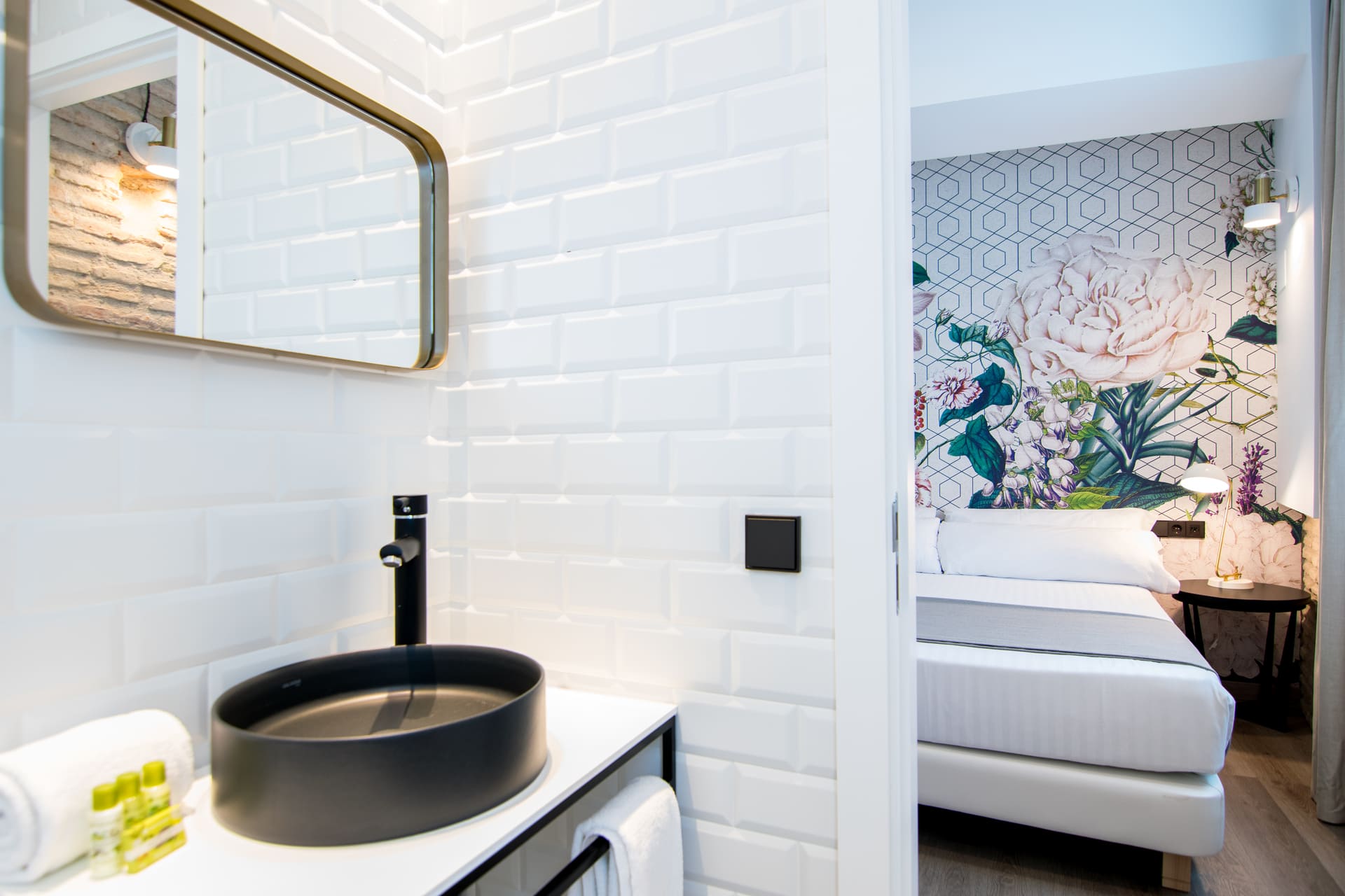 Lavabo negro y grifo en baño con azulejos blancos; dormitorio al fondo con cama y mural floral.
