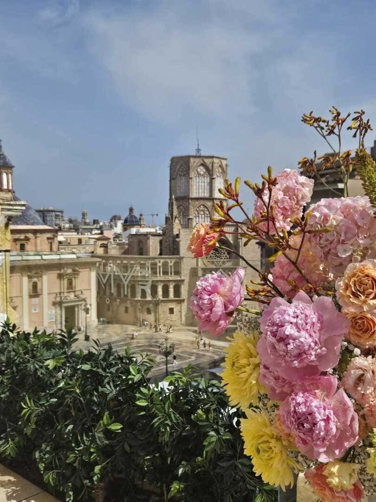 Plaza histórica con edificios antiguos y torre gótica, flores rosadas en primer plano.