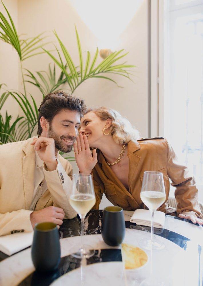Pareja sonriendo y susurrando junto a la mesa con copas de vino blanco