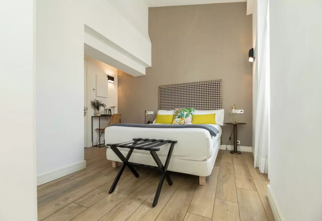 Habitación doble pequeña en Marques House con cama blanca, cabecero estampado y cojines amarillos.