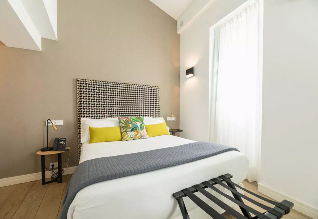 Habitación de hotel con cama grande y cabecera tapizada, cojines amarillos y florales, mesita de noche y ventana con cortinas.