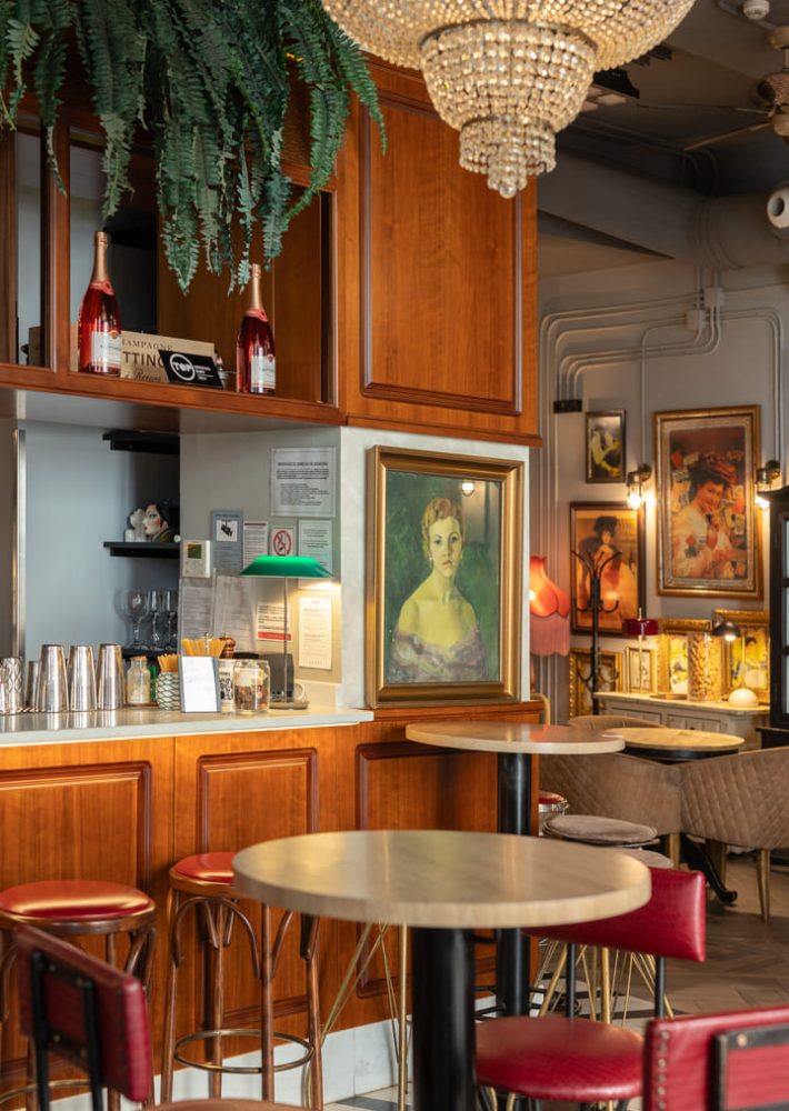 Bar elegante con bancone in legno, sgabelli rossi e lampadario, dipinti alle pareti e piante.