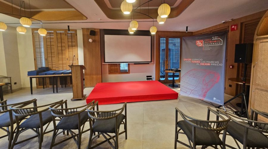 Escenario con plataforma roja en sala de conferencias, atril y pantalla grande, junto a un banner promocional y sillas para la audiencia.