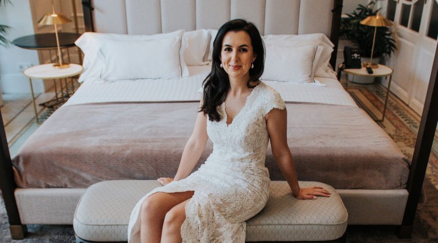 Novia con vestido de encaje blanco, sentada en banco en habitación de hotel