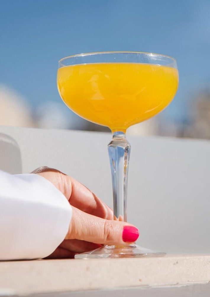 Cóctel naranja en copa coupe, sostenido por una mano en azotea con cielo azul