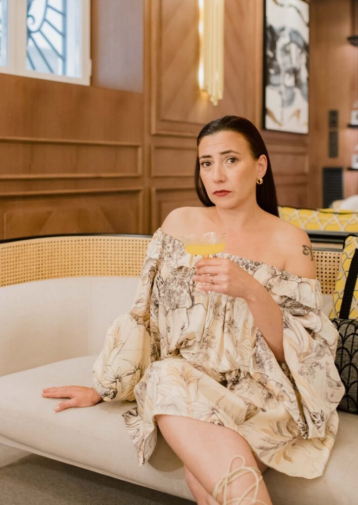 Mujer con vestido ligero de estampado floral y hombros descubiertos, sentada en sofá de lounge bar, sosteniendo un cóctel amarillo.