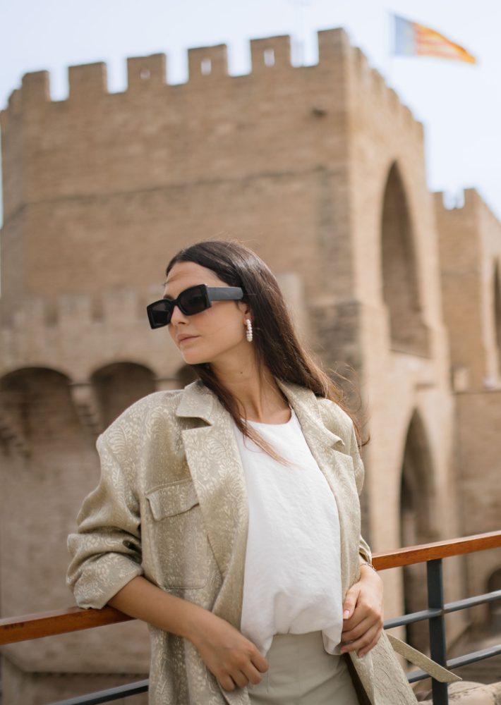 Mujer con blazer claro y gafas oscuras posando frente a murallas medievales