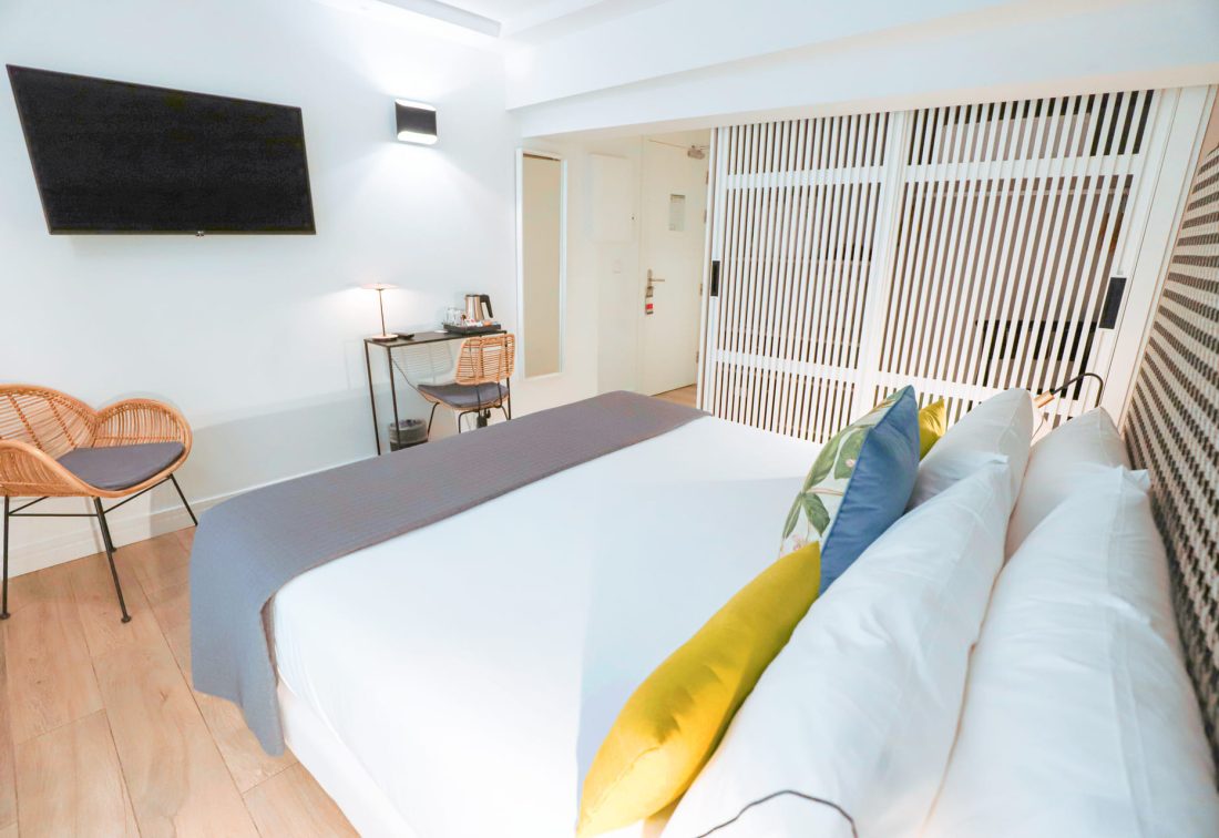 Habitación con cama grande, cojines multicolor, televisor y zona de trabajo con sillas de mimbre.