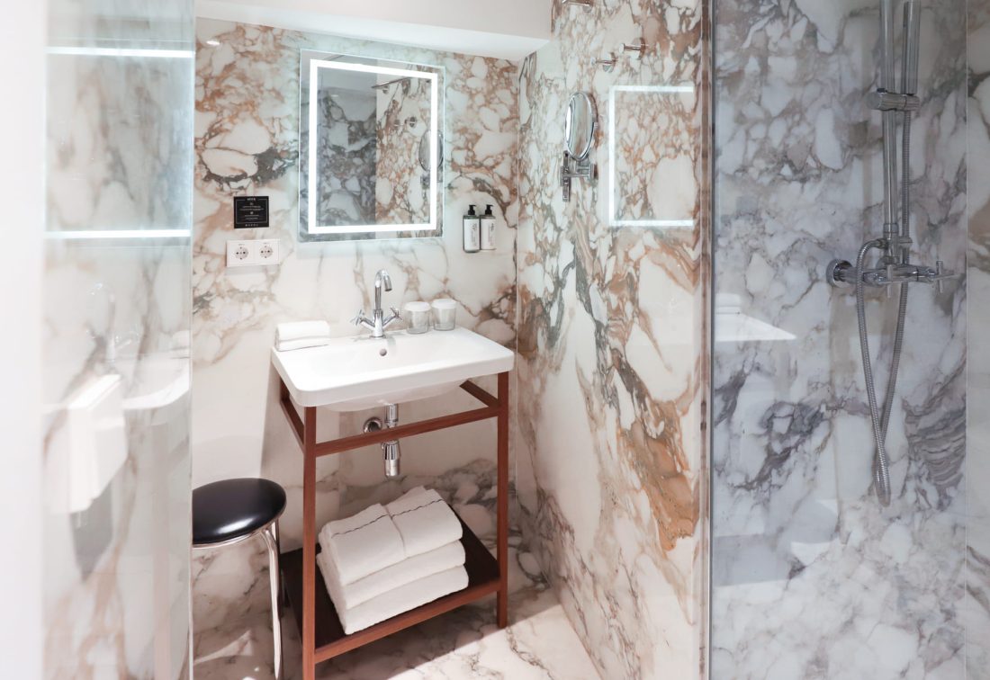 Baño de mármol con lavabo blanco sobre mueble de madera y ducha de vidrio