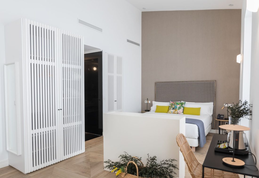 Habitación de hotel con cama y cabecero estampado, cojines verdes y zona de escritorio.