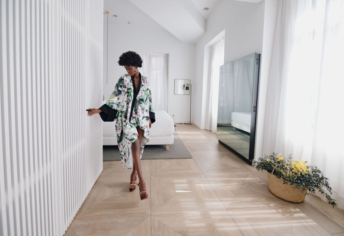 Mujer con kimono floral camina por pasillo de hotel, junto a cama blanca y espejo