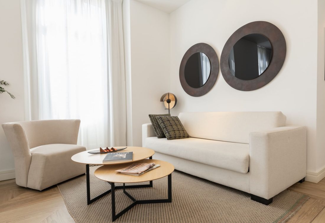 Sala de estar de habitación de hotel con sofá beige, sillón y dos espejos circulares en la pared