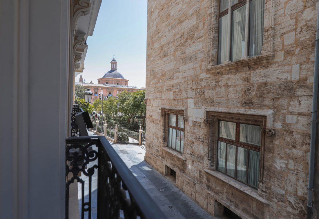 Balcón con barandilla de hierro forjado, vista hacia plaza con cúpula al fondo.