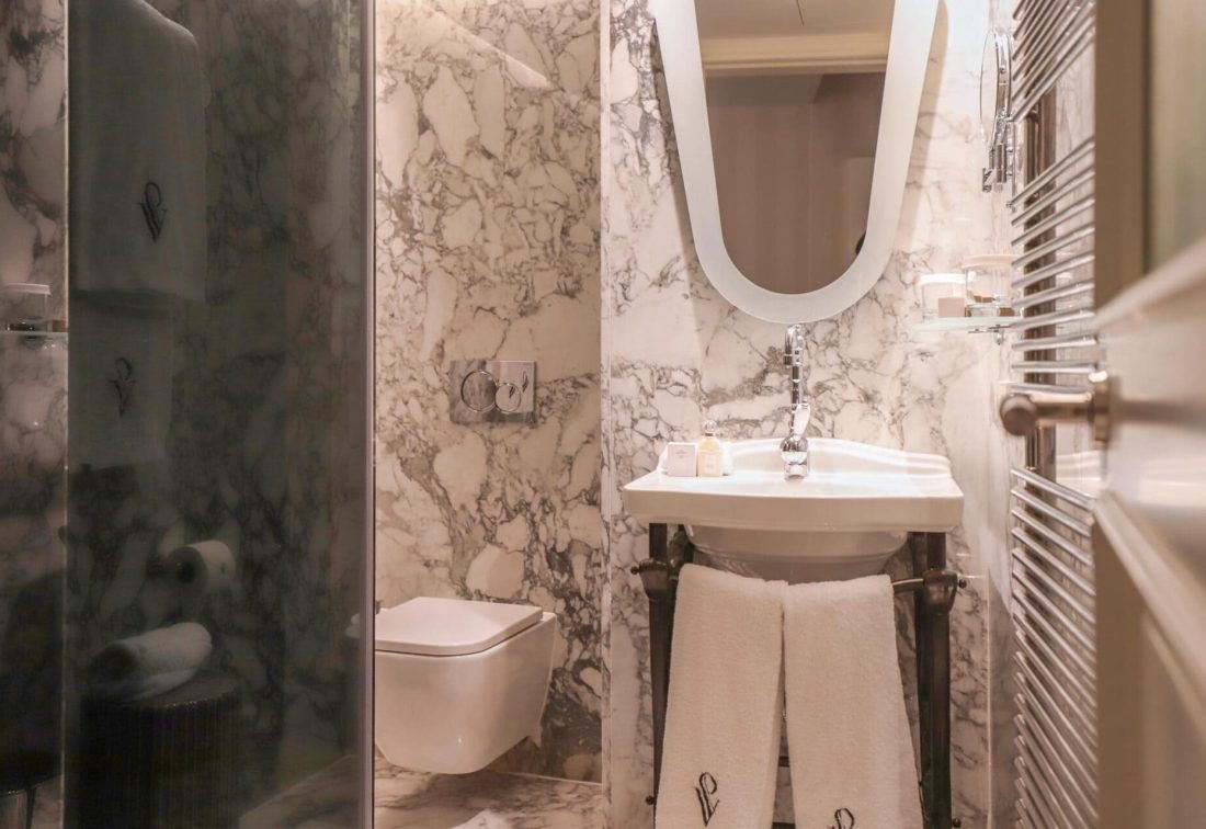 Baño de hotel con lavabo blanco, espejo ovalado, toallas colgando y paredes de mármol