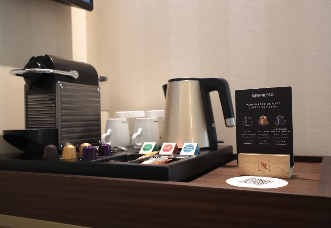 Estación de café en habitación con máquina Nespresso, hervidor y cápsulas.