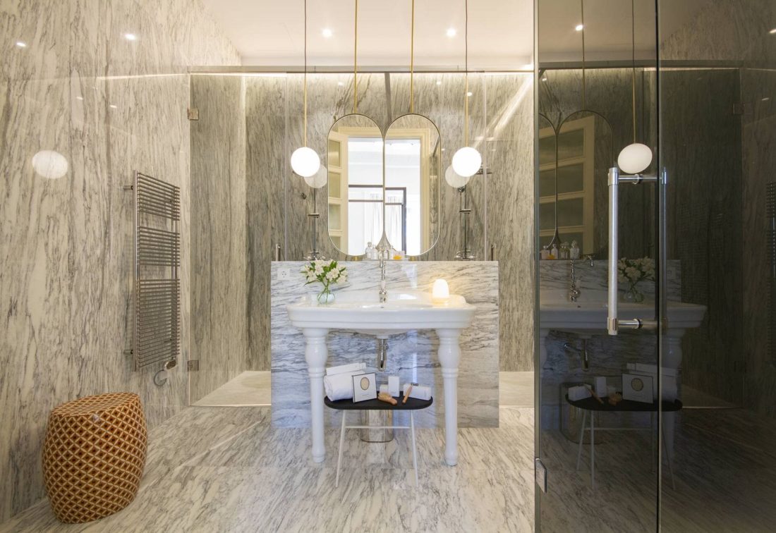 Baño de mármol con lavabo de pedestal blanco, espejo ovalado y ducha de vidrio; lámparas colgantes.