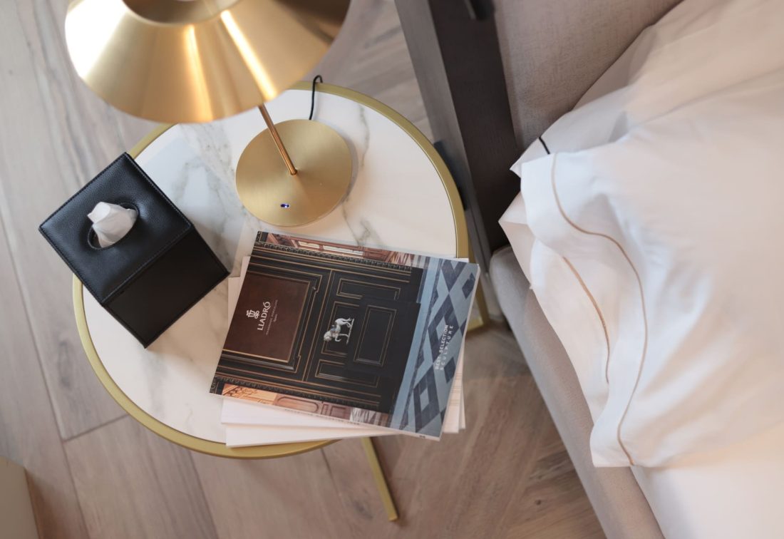 Mesa de noche redonda con superficie de mármol, lámpara dorada, caja de pañuelos y folleto junto a la cama.