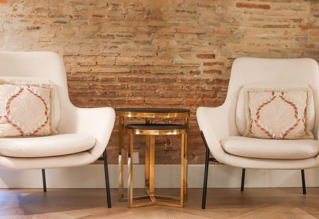 Dos sillones blancos con cojines decorativos entre ellos, junto a una mesa auxiliar dorada frente a una pared de ladrillo.