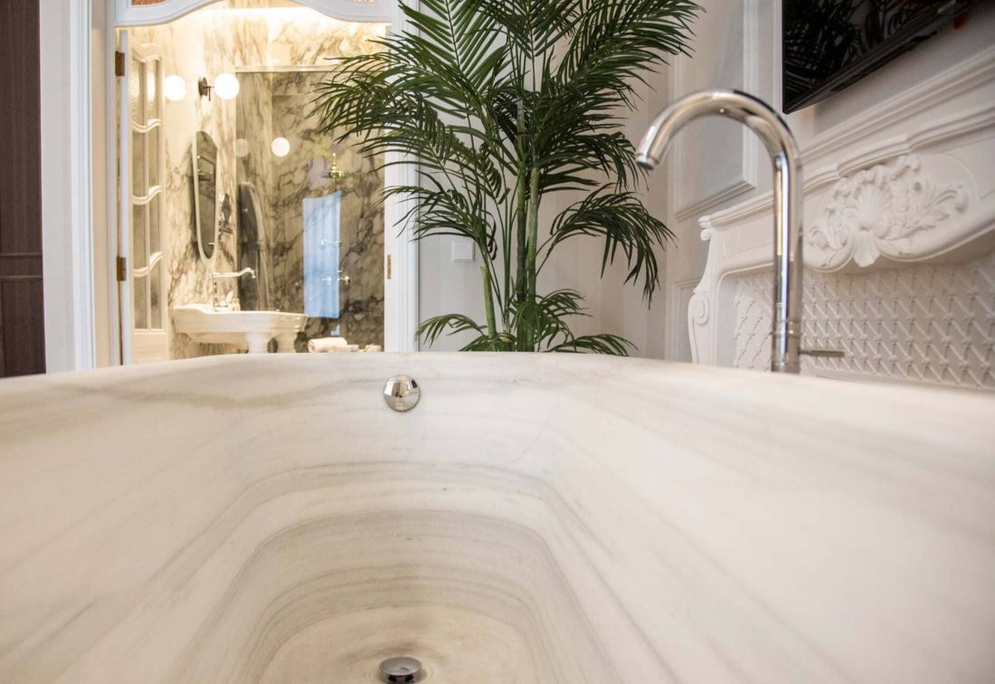 Bañera de mármol en baño de hotel, grifo de cuello alto y planta decorativa