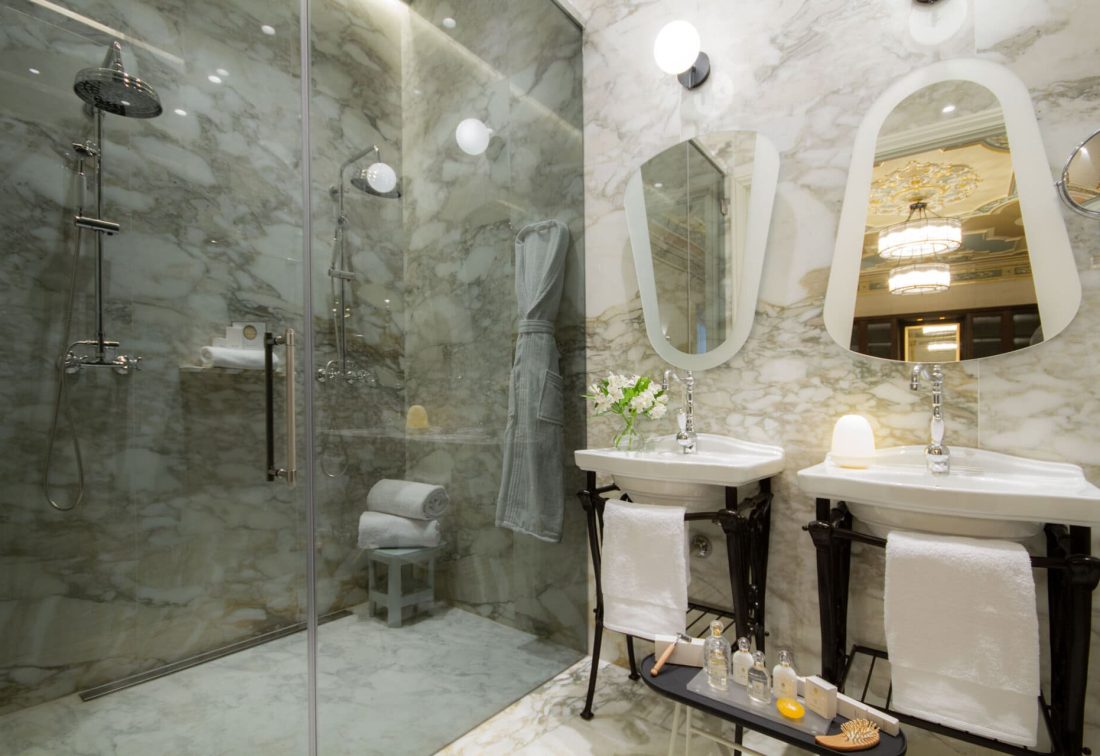 Baño de hotel con ducha de vidrio y lavabos dobles sobre mármol blanco