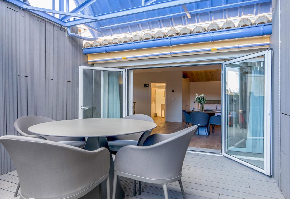 Terraza exterior con mesa redonda y sillas grises, puertas correderas abiertas a la sala de comedor.