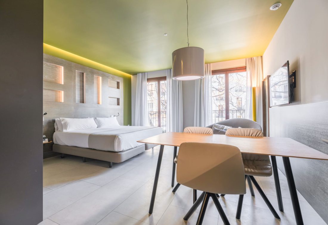 Habitación de hotel con cama doble, mesa de comedor y TV, junto a ventanales con cortinas.