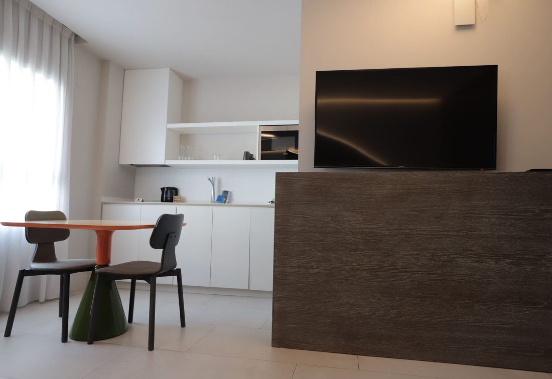 Sala de estar con cocina integrada, mesa redonda naranja y dos sillas, TV frente a panel de madera