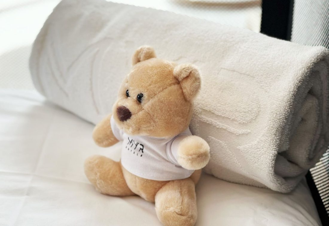 Oso de peluche con camiseta blanca en la cama junto a una toalla enrollada