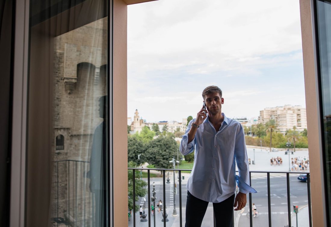 Hombre con camisa azul habla por teléfono en balcón de hotel con vista urbana