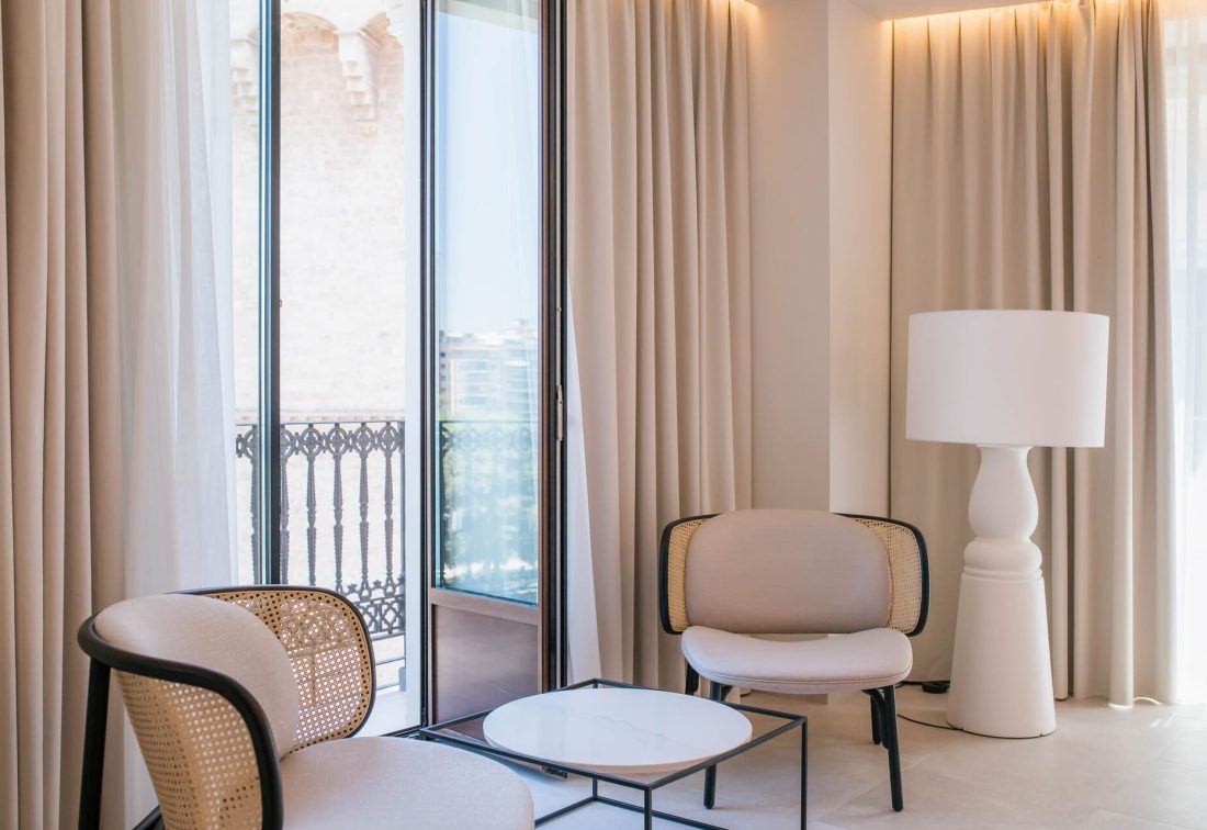 Rincón de descanso en habitación de hotel: dos sillones beige y mimbre, mesa redonda y lámpara junto a ventanales y balcón.