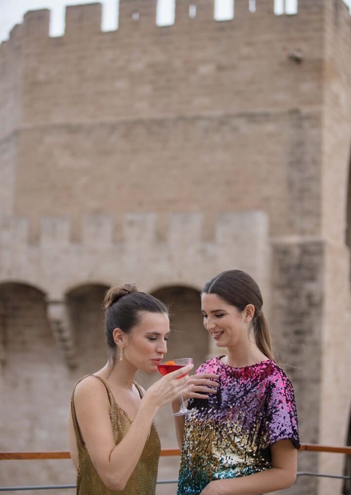 Dos mujeres con cócteles en Café Madrid Rooftop, frente a murallas históricas.