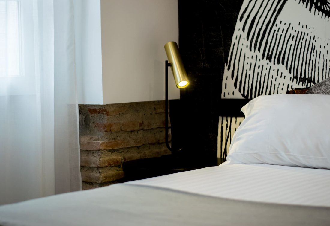 Cama de hotel con sábanas blancas junto a lámpara dorada y ventana con cortinas