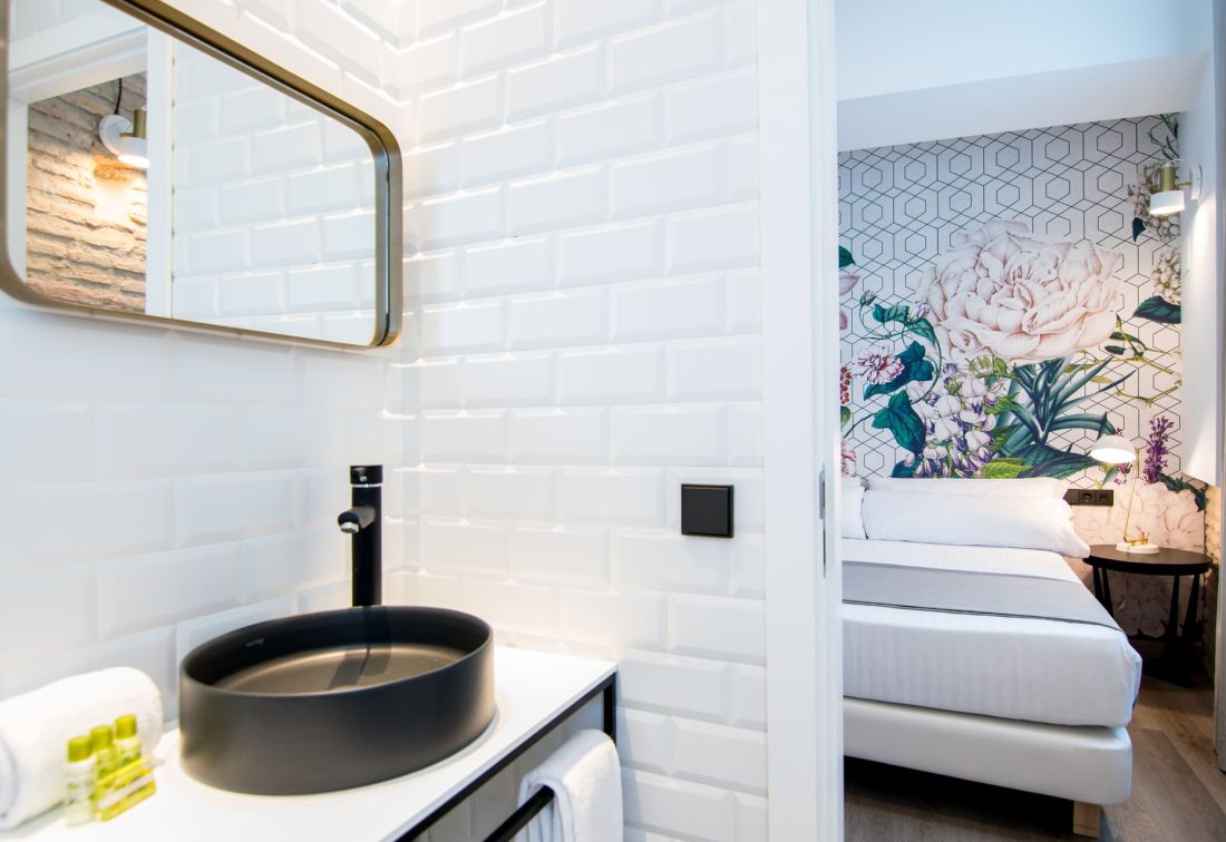 Lavabo negro y grifo en baño con azulejos blancos; dormitorio al fondo con cama y mural floral.
